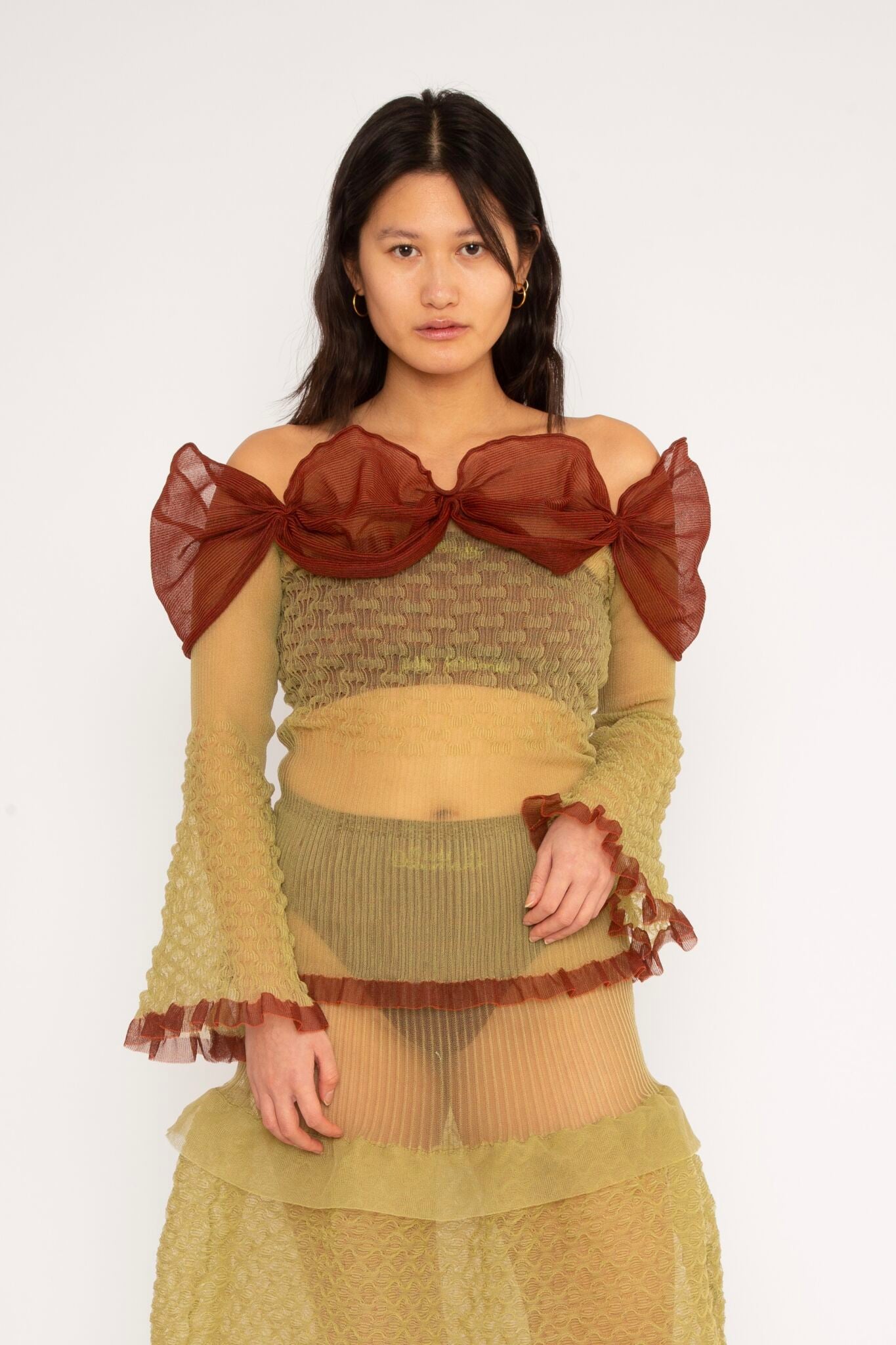 Bubble Bell Top in khaki and cherry red is a transparent knitted off-shoulder top in a ribbed bubble texture with ruffles and bell sleeves. The top has a close fit body with long sleeves and a voluminous adjustable neckline detailed with a melange rim with a logo in the back. The textile is lightweight, delicate and very flexible, meaning the dress adapts to body wearing it. The Bubble Bell Top is also available in teal and comes in 2 different sizes.