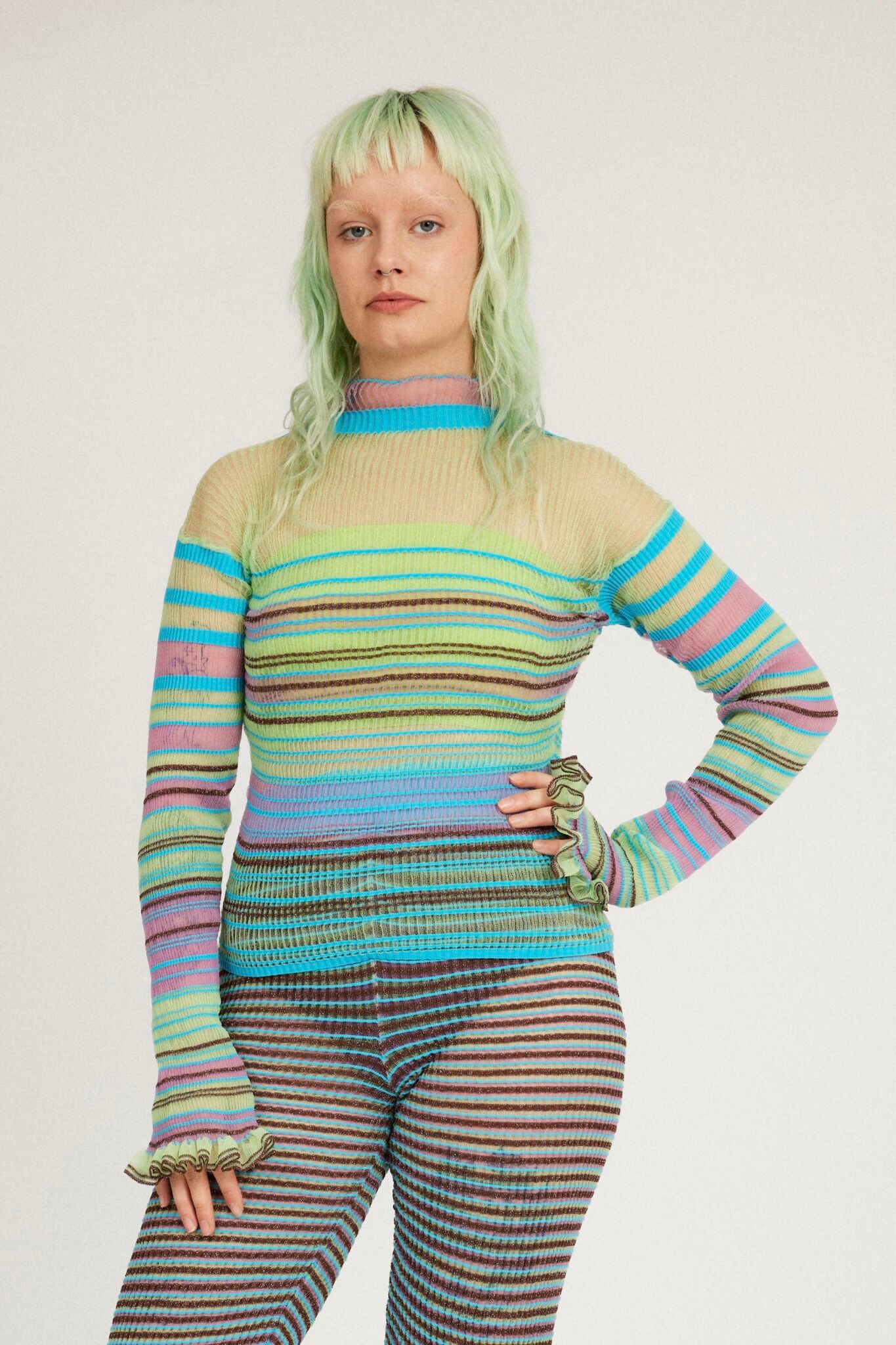 Sweet Secret Jumper in green and turquoise is a knitted transparent high neck jumper in all over bold stripes detailed with shimmery metallized yarn. The textile is lightweight, delicate and very flexible. The jumper has a body fitted silhouette that adapts to the body. Detailed with frills and a logo in the back of the neck.