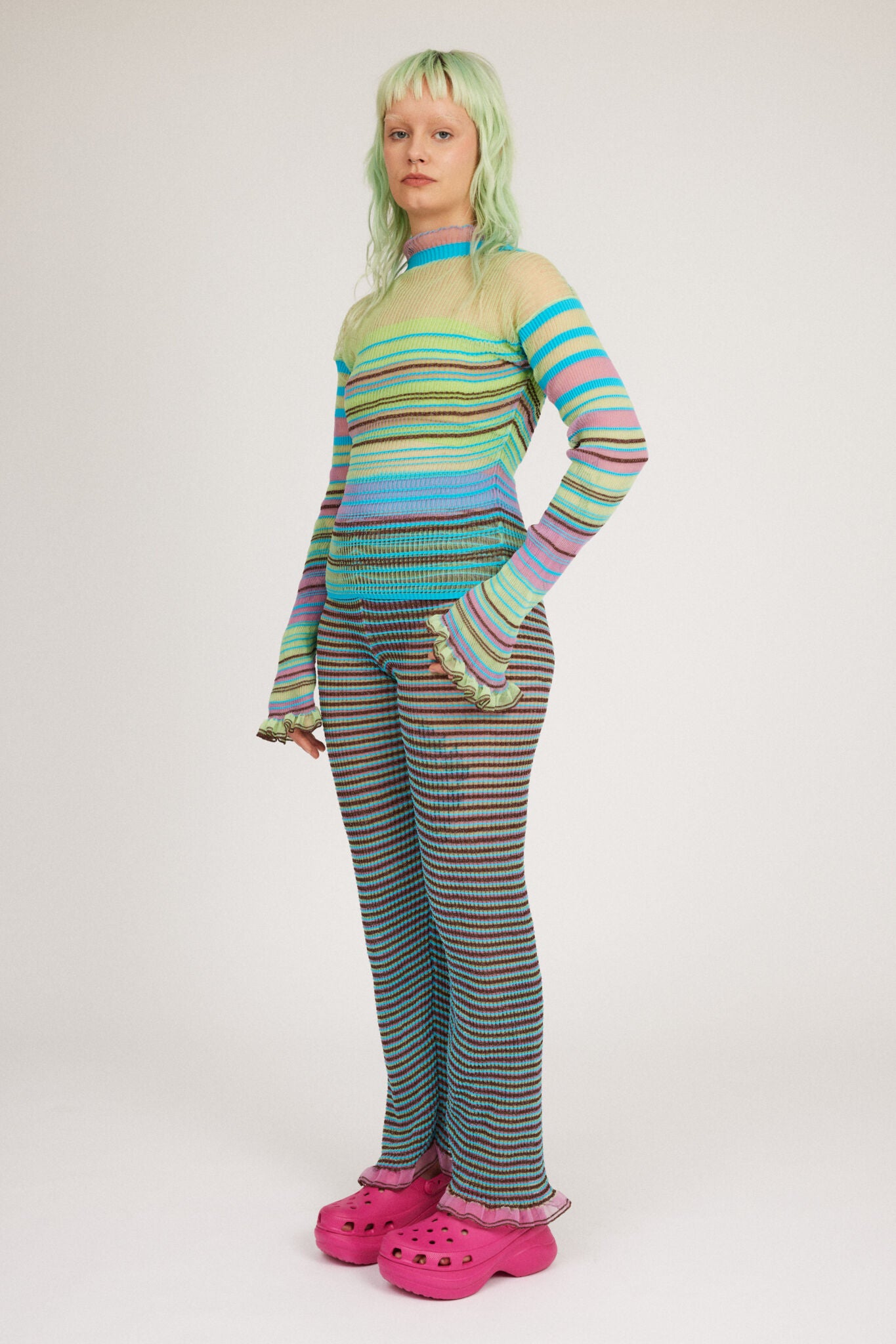 Sweet Secret Jumper in green and turquoise is a knitted transparent high neck jumper in all over bold stripes detailed with shimmery metallized yarn. The textile is lightweight, delicate and very flexible. The jumper has a body fitted silhouette that adapts to the body. Detailed with frills and a logo in the back of the neck.
