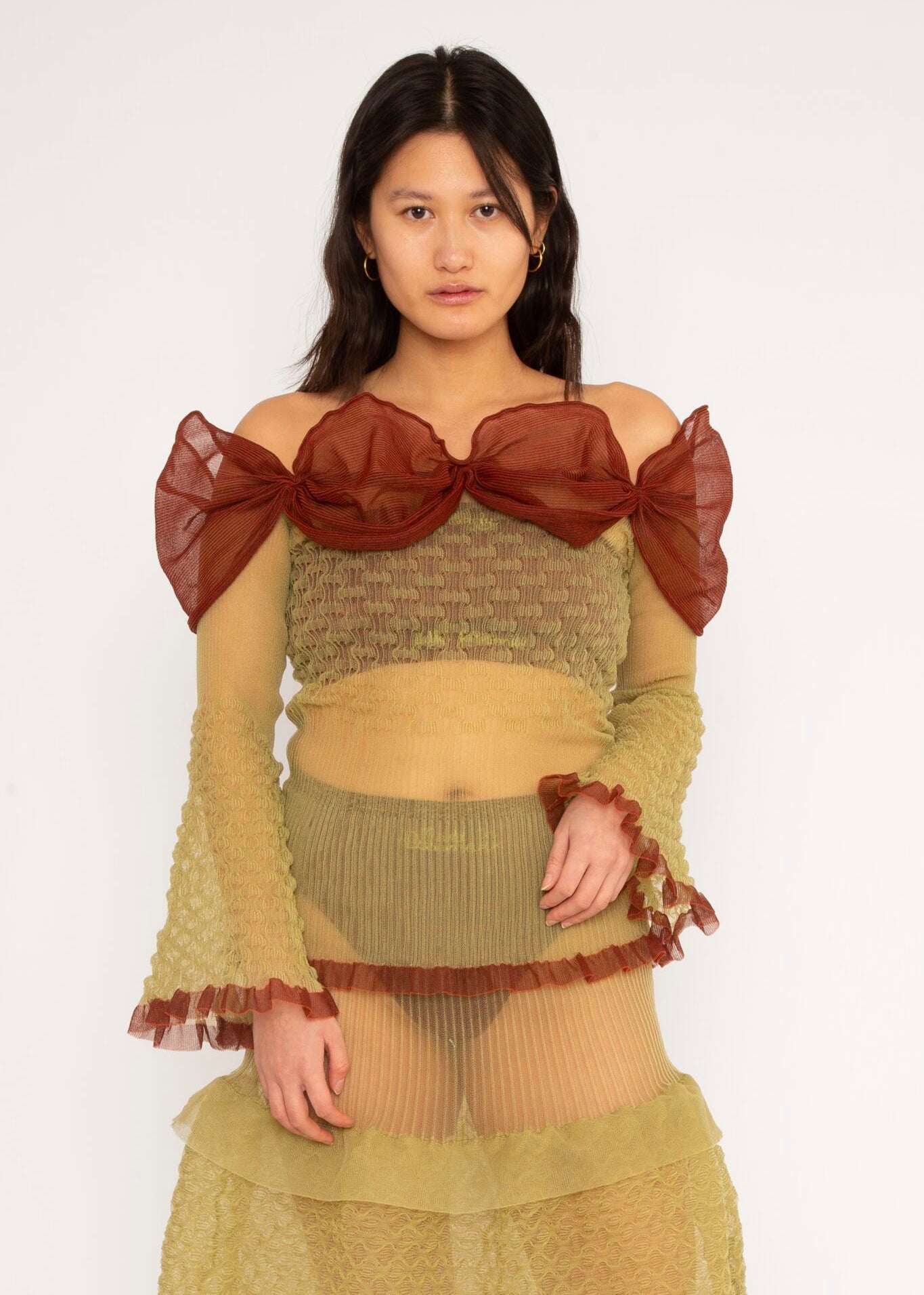 Bubble Bell Top in khaki and cherry red is a transparent knitted off-shoulder top in a ribbed bubble texture with ruffles and bell sleeves. The top has a close fit body with long sleeves and a voluminous adjustable neckline detailed with a melange rim with a logo in the back. The textile is lightweight, delicate and very flexible, meaning the dress adapts to body wearing it. The Bubble Bell Top is also available in teal and comes in 2 different sizes.