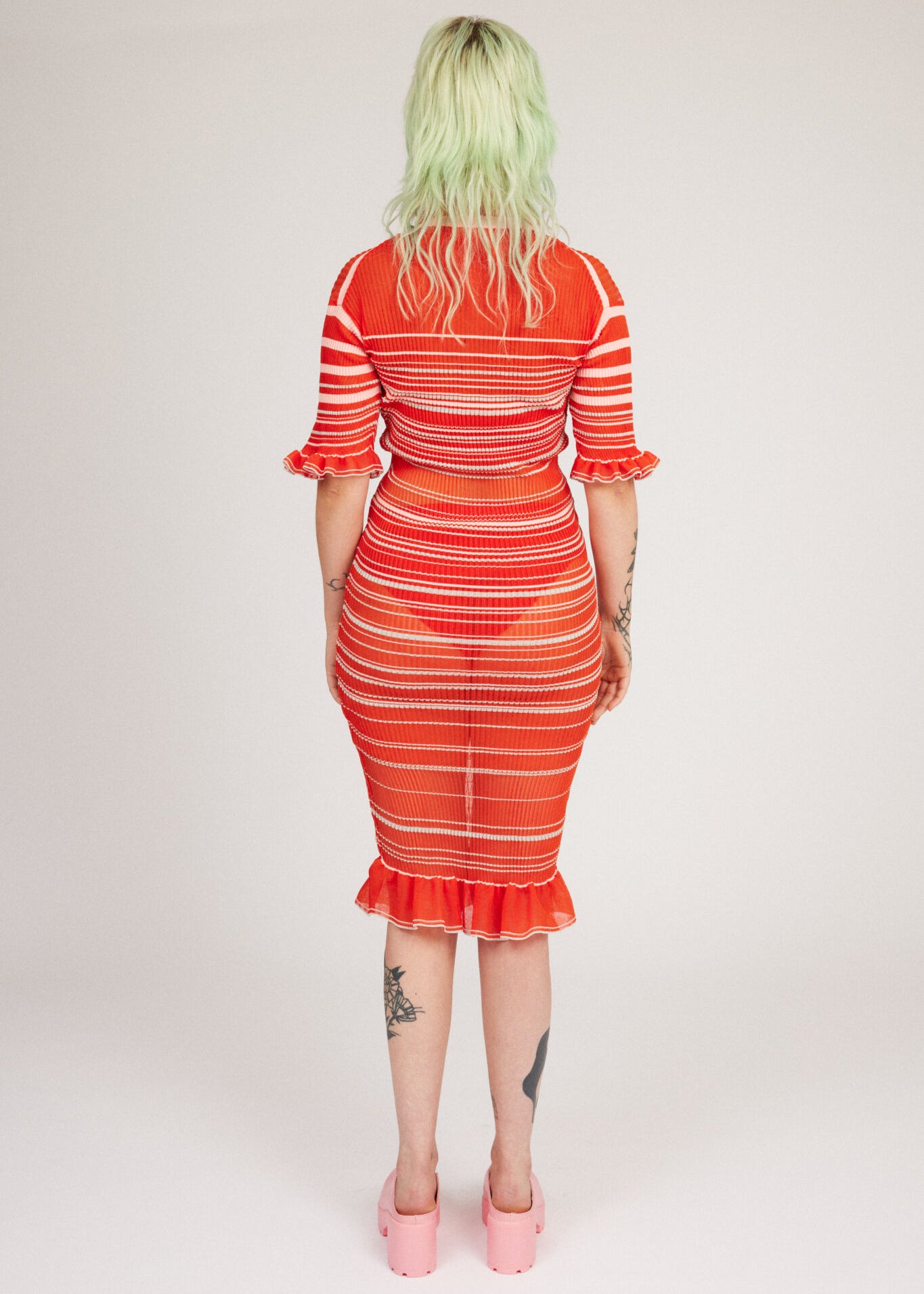 Sweet Secret Dress in coral red is a transparent knitted short sleeved high neck dress in gradient stripes with shimmery metallized yarn. The textile is lightweight, delicate and very flexible. The dress has a body fitted silhouette that adapts to the body. Detailed with frills and a logo in the back of neck.