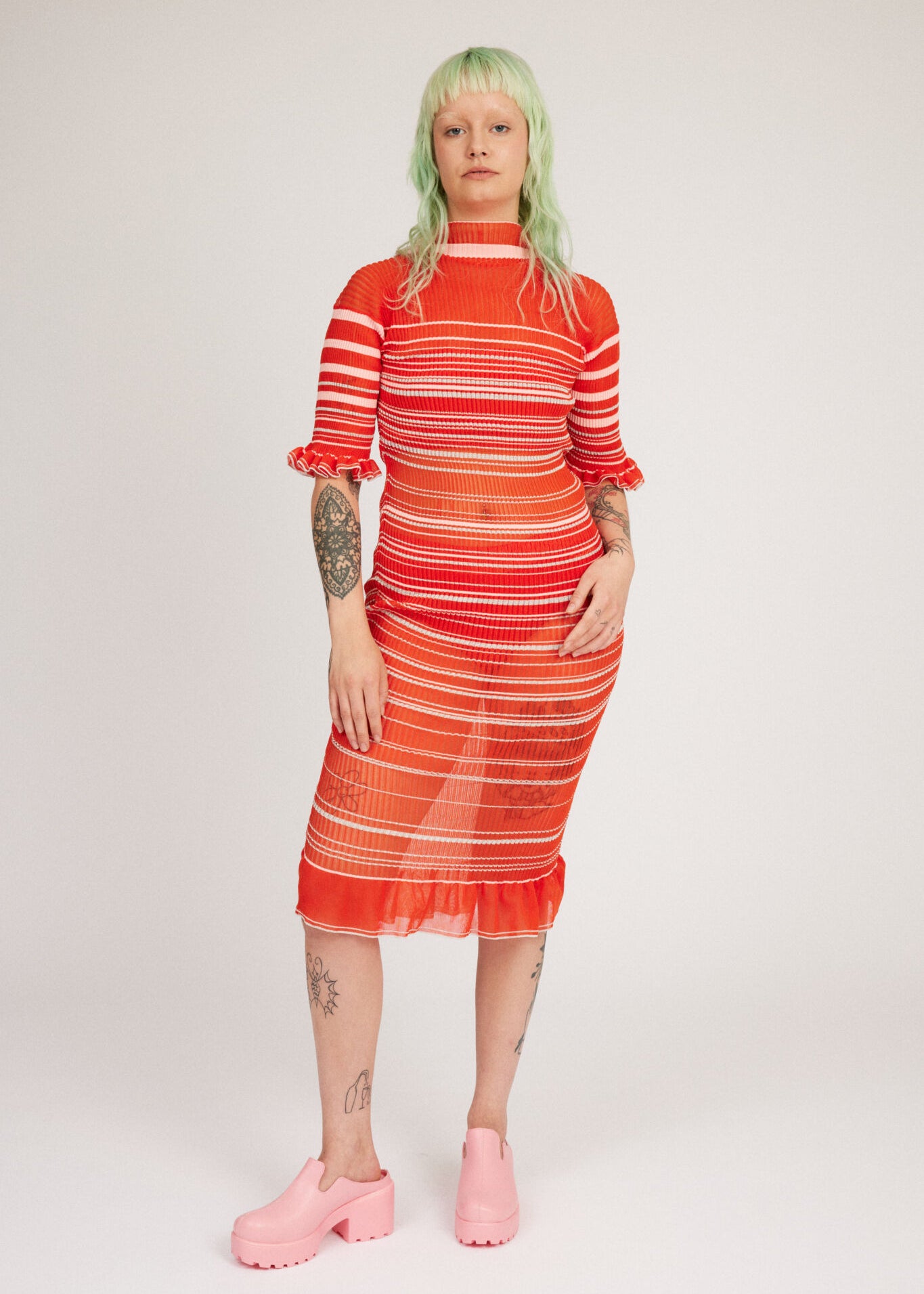 Sweet Secret Dress in coral red is a transparent knitted short sleeved high neck dress in gradient stripes with shimmery metallized yarn. The textile is lightweight, delicate and very flexible. The dress has a body fitted silhouette that adapts to the body. Detailed with frills and a logo in the back of neck.