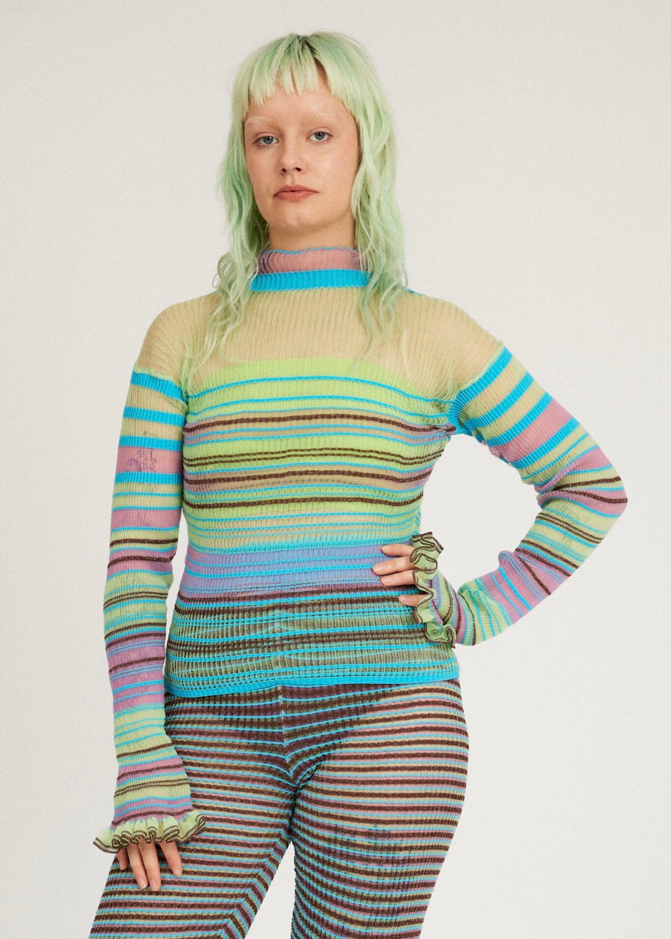 Sweet Secret Jumper in green and turquoise is a knitted transparent high neck jumper in all over bold stripes detailed with shimmery metallized yarn. The textile is lightweight, delicate and very flexible. The jumper has a body fitted silhouette that adapts to the body. Detailed with frills and a logo in the back of the neck.