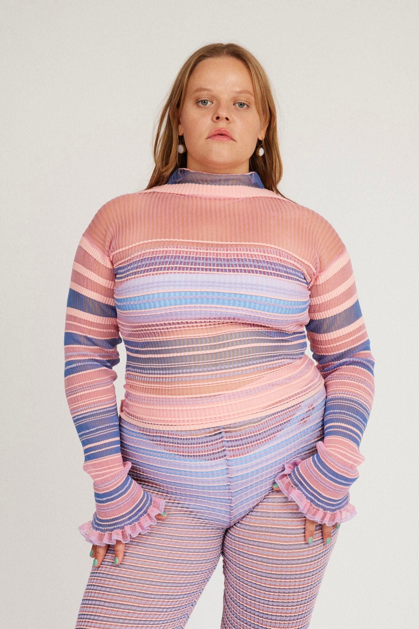 Sweet Secret Jumper in pink and powder blue is a knitted transparent high neck jumper in all over bold stripes detailed with shimmery metallized yarn. The textile is lightweight, delicate and very flexible. The jumper has a body fitted silhouette that adapts to the body. Detailed with frills and a logo in the back of the neck.
