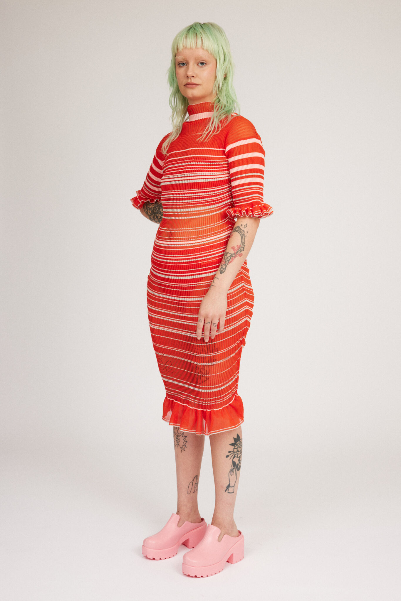 Sweet Secret Dress in coral red is a transparent knitted short sleeved high neck dress in gradient stripes with shimmery metallized yarn. The textile is lightweight, delicate and very flexible. The dress has a body fitted silhouette that adapts to the body. Detailed with frills and a logo in the back of neck.