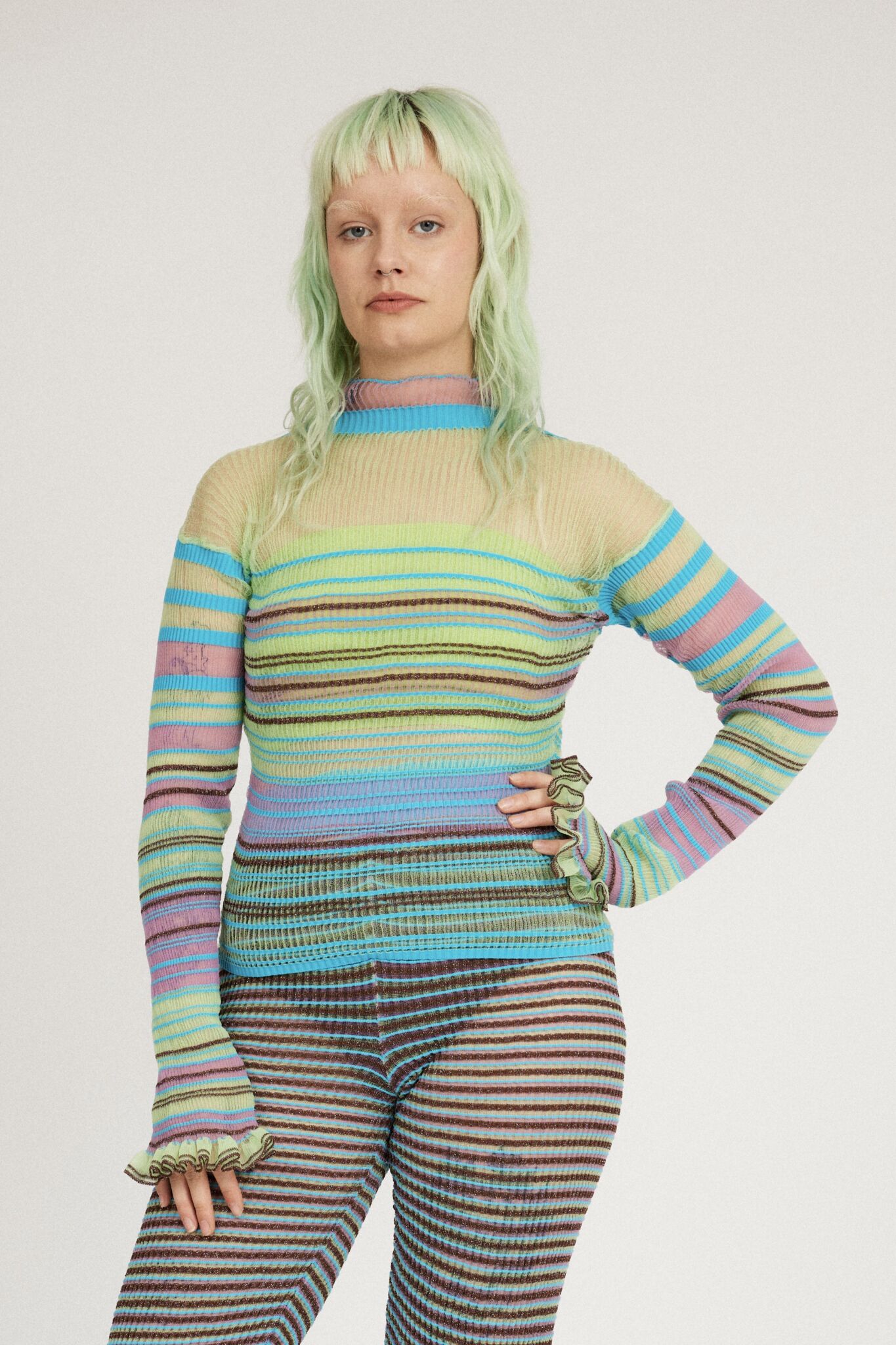 Sweet Secret Jumper in green and turquoise is a knitted transparent high neck jumper in all over bold stripes detailed with shimmery metallized yarn. The textile is lightweight, delicate and very flexible. The jumper has a body fitted silhouette that adapts to the body. Detailed with frills and a logo in the back of the neck.