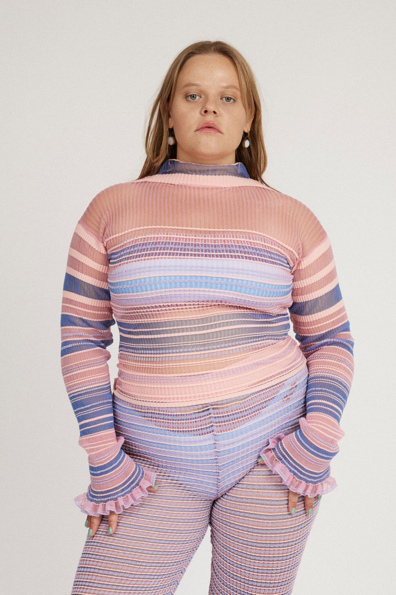Sweet Secret Jumper in pink and powder blue is a knitted transparent high neck jumper in all over bold stripes detailed with shimmery metallized yarn. The textile is lightweight, delicate and very flexible. The jumper has a body fitted silhouette that adapts to the body. Detailed with frills and a logo in the back of the neck.
