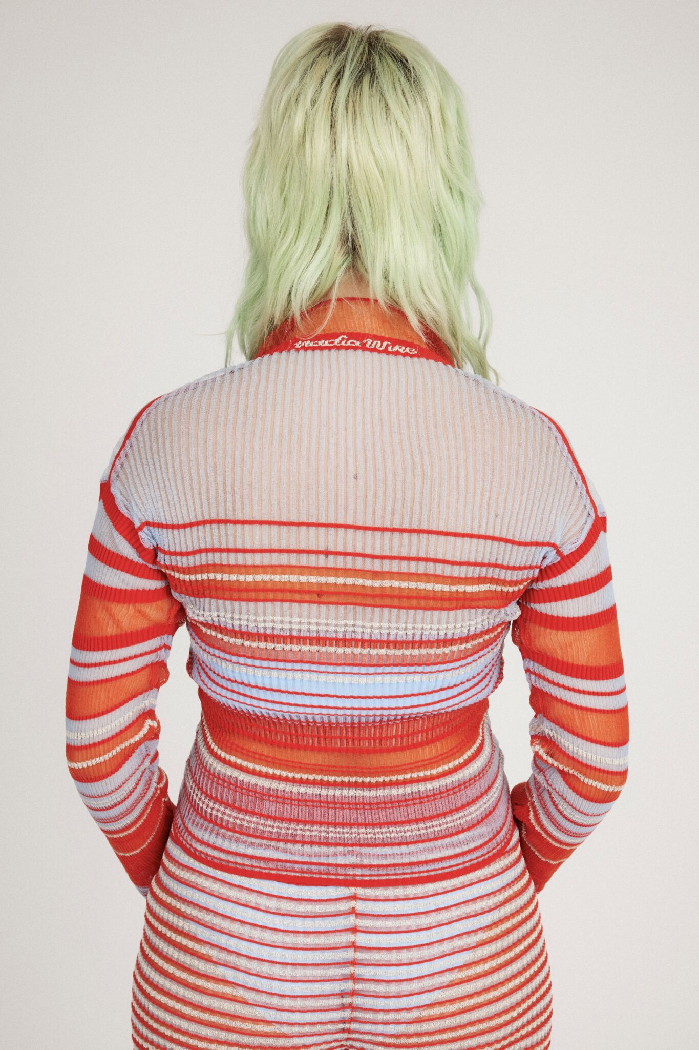 Sweet Secret Jumper in coral red and light blue is a knitted transparent high neck jumper in all over bold stripes detailed with shimmery metallized yarn. The textile is lightweight, delicate and very flexible. The jumper has a body fitted silhouette that adapts to the body. Detailed with frills and a logo in the back of the neck.