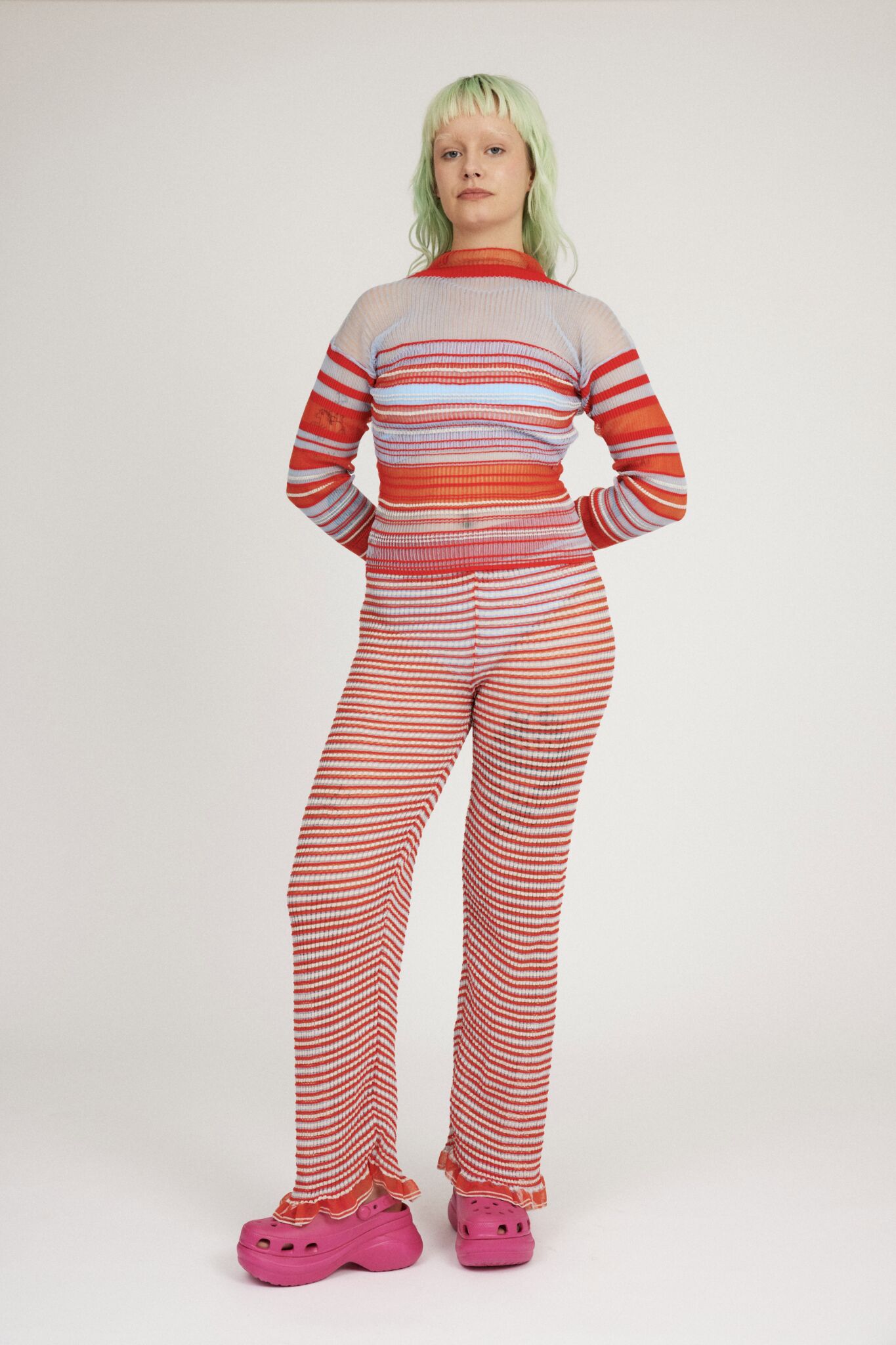 Sweet Secret Trouser in coral red and light blue are a knitted pair of transparent trousers in all over stripes with shimmery metallized yarn. The textile is lightweight, delicate and very flexible. The trousers have a body fitted silhouette that adapts to the body. Wide legged and detailed with frills and a logo waistband.