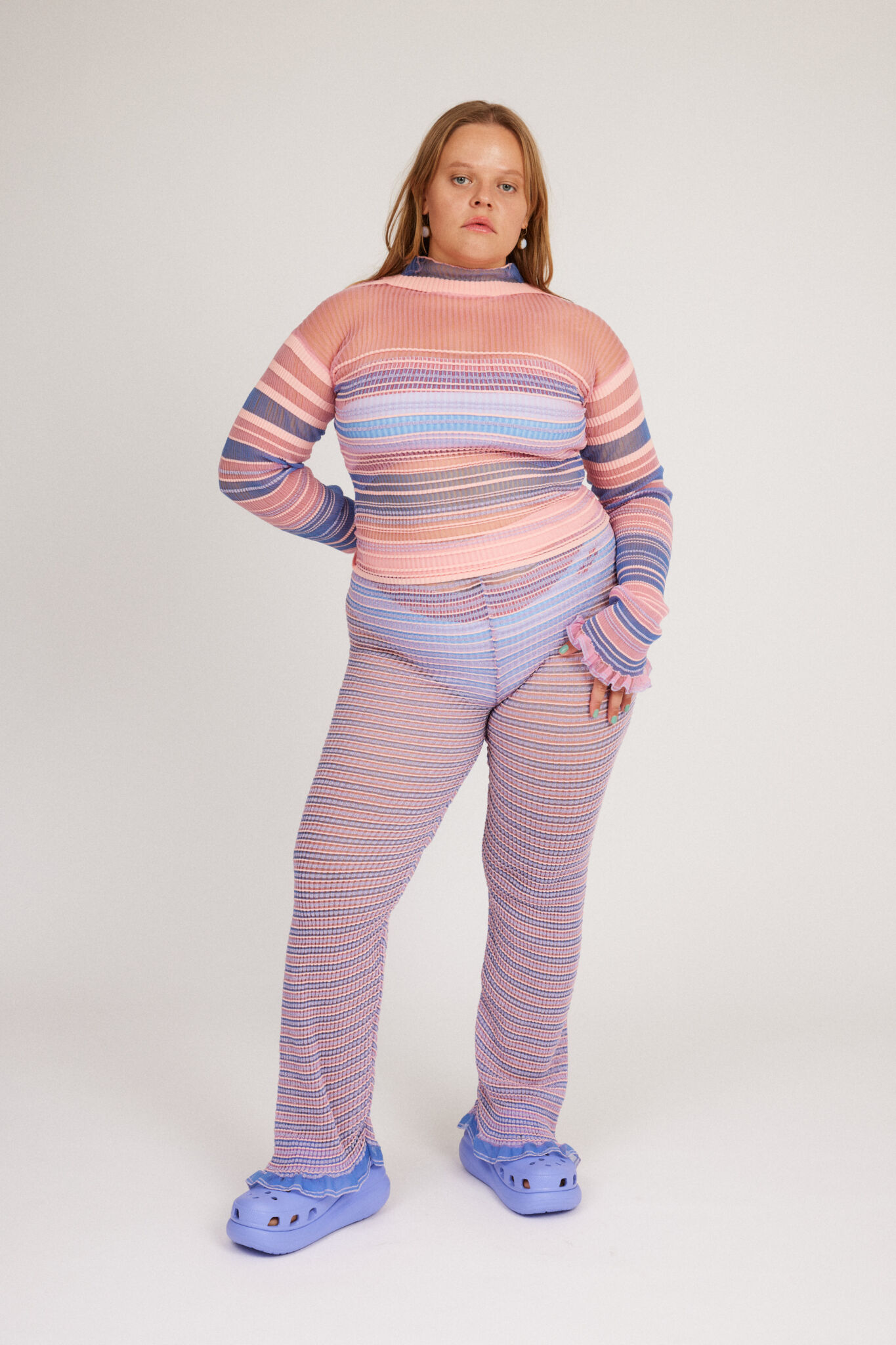 Sweet Secret Trouser in pink and powder blue are a knitted pair of transparent trousers in all over stripes with shimmery metallized yarn. The textile is lightweight, delicate and very flexible. The trousers have a body fitted silhouette that adapts to the body. Wide legged and detailed with frills and a logo waistband.