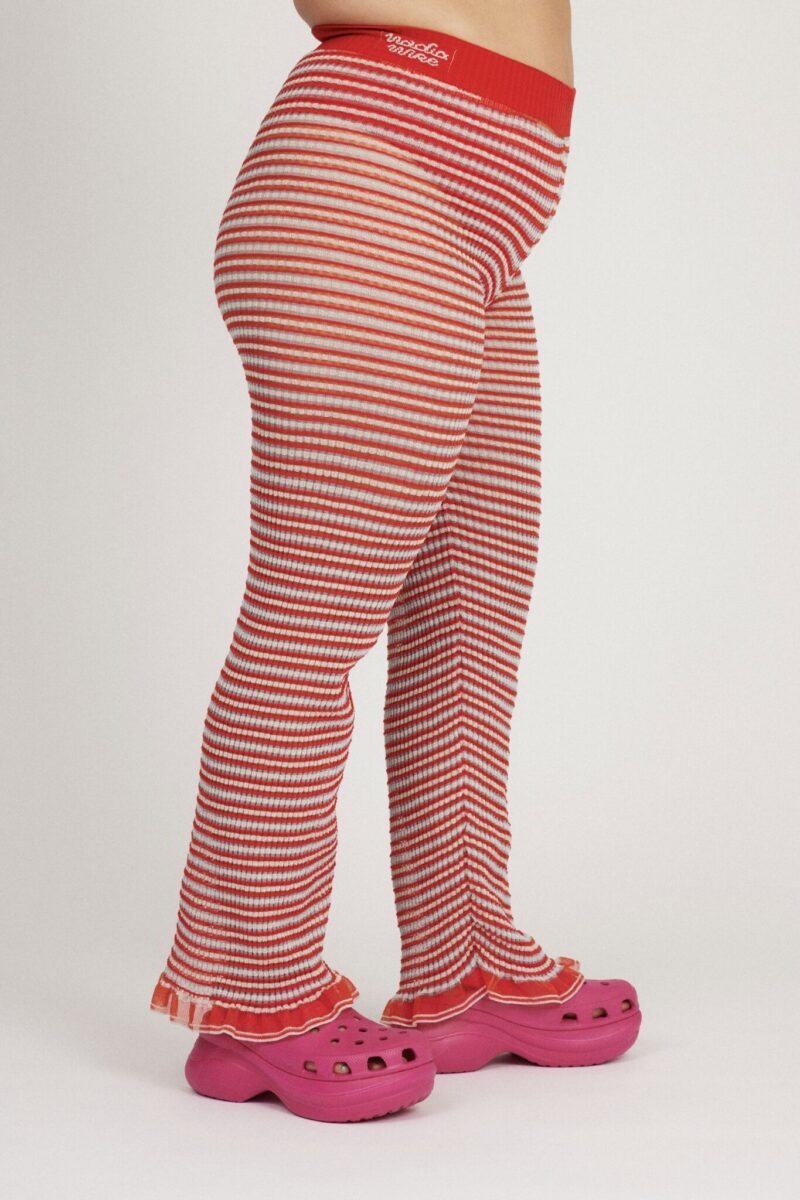 Sweet Secret Trouser in coral red and light blue are a knitted pair of transparent trousers in all over stripes with shimmery metallized yarn. The textile is lightweight, delicate and very flexible. The trousers have a body fitted silhouette that adapts to the body. Wide legged and detailed with frills and a logo waistband.