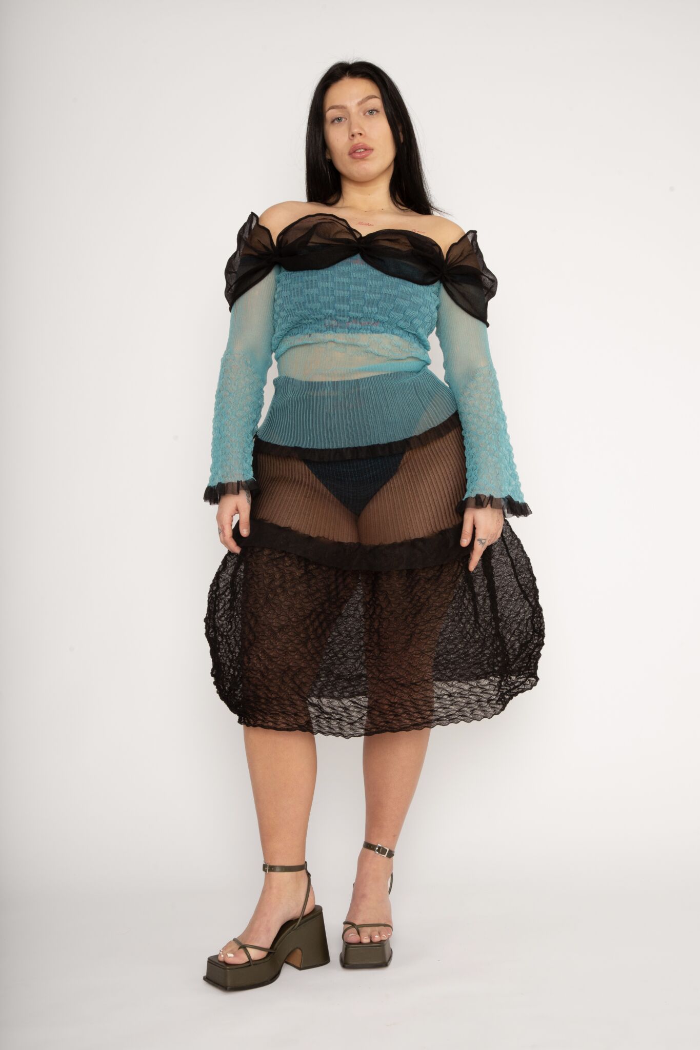 Bubble Bell Top in teal and black is a transparent knitted off-shoulder top in a ribbed bubble texture with ruffles and bell sleeves. The top has a close fit body with long sleeves and a voluminous adjustable neckline detailed with a melange rim with a logo in the back. The textile is lightweight, delicate and very flexible, meaning the dress adapts to body wearing it. The Bubble Bell Top is also available in khaki and comes in 2 different sizes.