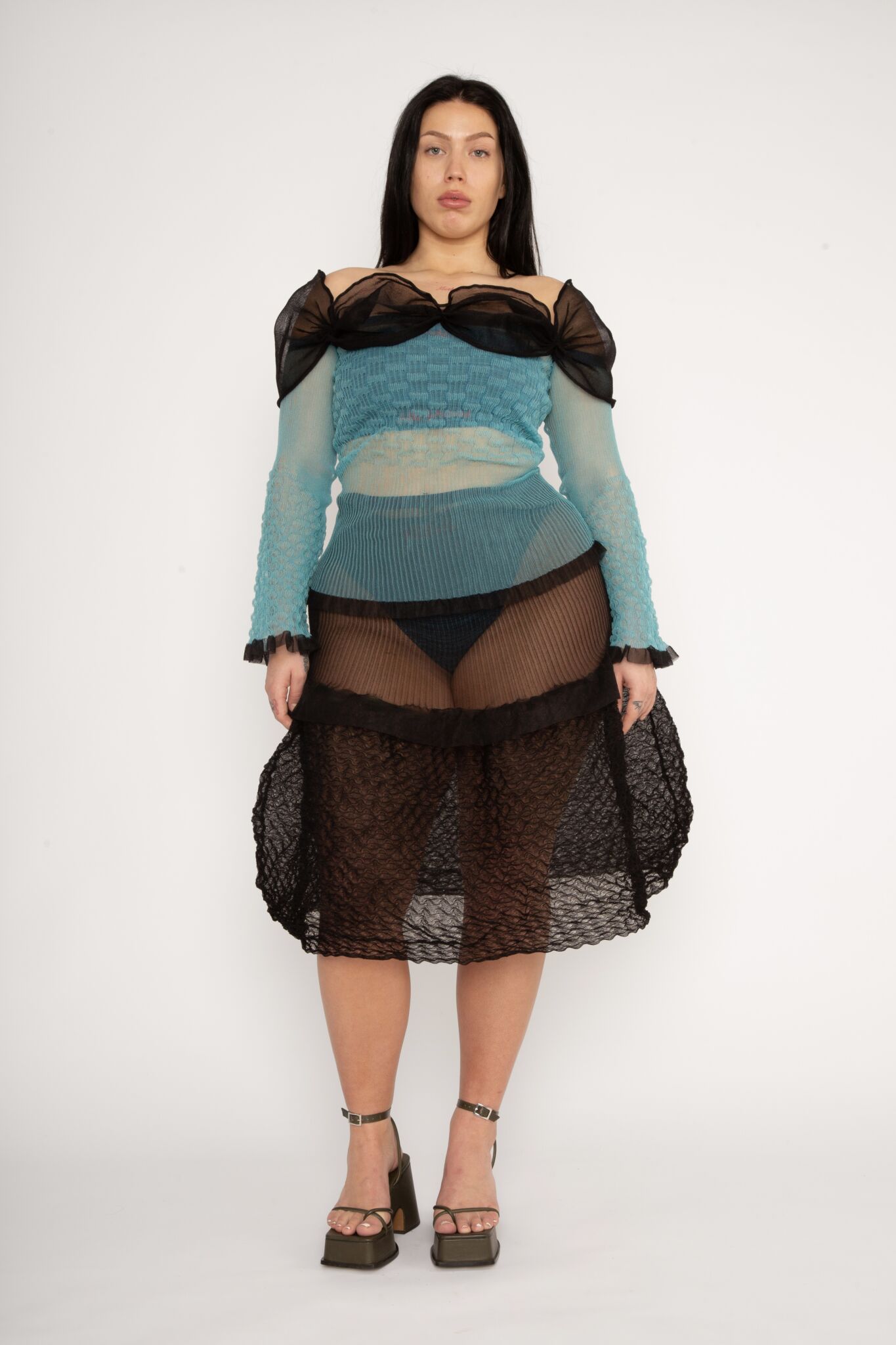 Bubble Bell Top in teal and black is a transparent knitted off-shoulder top in a ribbed bubble texture with ruffles and bell sleeves. The top has a close fit body with long sleeves and a voluminous adjustable neckline detailed with a melange rim with a logo in the back. The textile is lightweight, delicate and very flexible, meaning the dress adapts to body wearing it. The Bubble Bell Top is also available in khaki and comes in 2 different sizes.